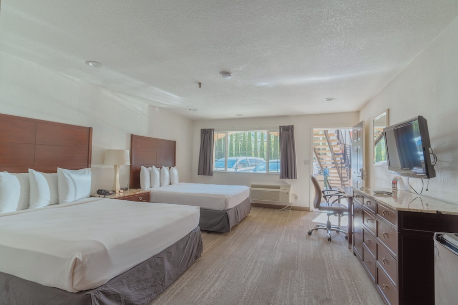 A hotel room with two beds, a desk, a TV, and a window with a view of buildings and cars. - The Yosemite Inn (Mariposa)